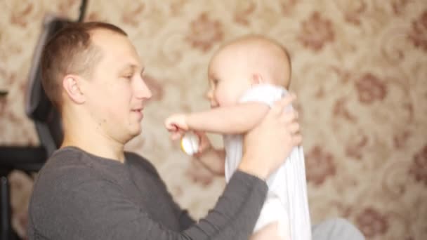 Vader, zit op de vloer en houdt zijn baby in de handen, speelt met zijn zoon, kust hem en ze lachen allebei gelukkig. Zijaanzicht medium schot in 4k video. — Stockvideo