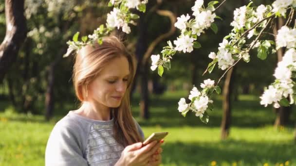 Mujer bonita feliz está sonriendo y mensajes de texto en el teléfono inteligente en el fondo de un manzano en flor en el parque de la ciudad de primavera. Concepto de mujer retrato fotografía, gadgets o viajar. 4K video en cámara lenta . — Vídeo de stock