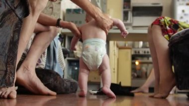 Dedem bebeği kollarının altında destekliyor yere ilk basamaklarını atmasına yardım ediyor ve büyük sandalyede oturan büyükannesi de mutlu bir şekilde gülümsüyor. 4k video 'da alt görünüm orta görüntüsü.