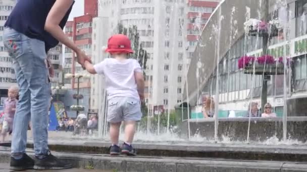 Fıskiyenin yanında bebeğini tutan genç bir kadın oğluna arka planda şehir merkeziyle akan suyu gösteriyor. 4k yavaş çekim geri görüntü videosu. — Stok video