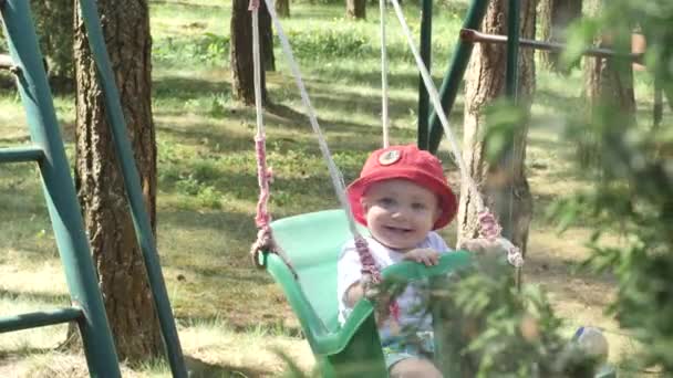 Heureux sourire petit bébé en panama rouge avec des piqûres de moustiques sur son visage est monté sur une balançoire dans le parc avec des arbres sur le fond, puis sa mère vient à lui au ralenti vue de côté 4K vidéo . — Video