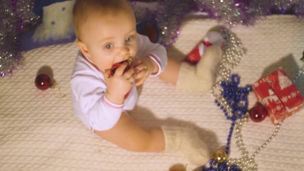 De kleine jongen zit op de vloer bij de versierde kerstboom en bijt de rode kerstbal in zijn handen en kijkt in de camera. Vrolijk kerst- en nieuwjaarsconcept in 4k slow motion top view video. — Stockvideo