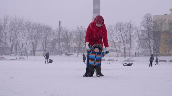 Mujer joven en chaqueta de plumón rojo ayuda a su bebé a caminar bajo la nieve a la cámara en el fondo del parque de la ciudad de invierno en cámara lenta de medio disparo 4K video . — Foto de Stock