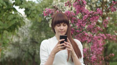 Kız bahçede selfie çekiyor. Çekici kızıl saçlı bir kadın kiraz bahçesinde cep telefonu kullanarak selfi yaparken gülümsüyor. Cihazları sağlıklı bir yaşam tarzı için kullanma kavramı.