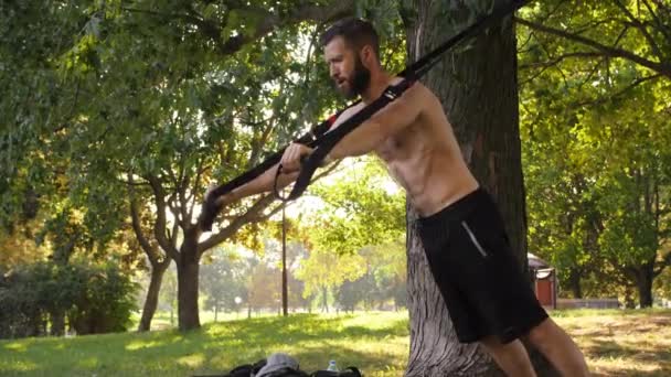 Barbuto giovane uomo muscoloso fare esercizio con cicli di fitness vicino a un albero. 4K lento mo primo piano filmato — Video Stock