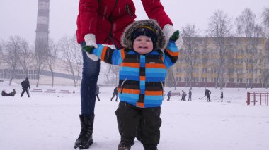 Anne, parktaki karda gülümseyen küçük bir bebeğin ellerine önderlik ediyor. Çocuğun ilk adımları. Ebeveyn bakımı