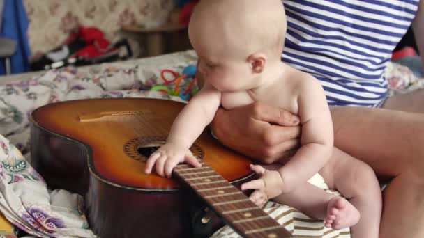 Grandfather and his toddler grandson are sitting on the sofa near a vintage acoustic guitar and the baby boy looks at instrument and touches strings. Family concept. Front view medium shot in 4K video — 비디오