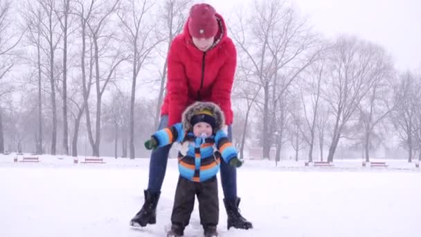 Giovane donna in piumino rosso rotola il suo bambino figlio su ghiaccio sotto la neve sullo sfondo del parco urbano invernale in slow motion medio girato video 4K . — Video Stock