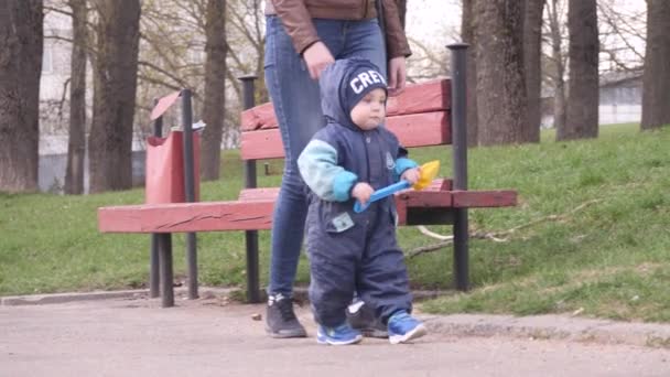 Young woman with her little baby boy who holding baby shovel are walking in the city park in a spring cloudy day and kid tries to run for a dove in 4K slow motion video. — 비디오