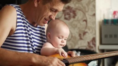 Büyükbaba küçük torununa klasik bir akustik gitar gösteriyor ve çocuk merakla ona bakıyor ve tellere dokunuyor. Mutlu aile kavramı. 4k video içinde kenar görünümü orta görüntüsü.