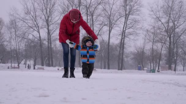 Jonge moeder in rood donsjas helpt haar kleine jongen om zijn eerste stappen te zetten en ze lopen samen naar de camera op de winter stadspark achtergrond in slow motion medium shot 4k video. — Stockvideo