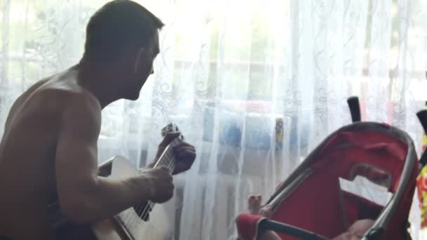 Ο γηραιότερος παίζει κιθάρα και τραγουδά για τον εγγονό του που ξαπλώνει στο κόκκινο καροτσάκι του μωρού στο φόντο του παραθύρου. Μεσαία προβολή πλαϊνής όψης σε βίντεο 4k. — Αρχείο Βίντεο