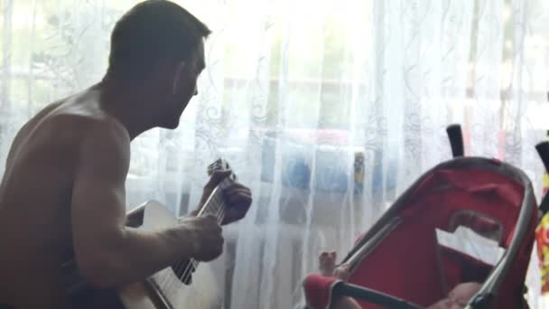 Elder man is playing on acoustic guitar and singing song for his little grandson who laying in red baby carriage at window background. Side view medium shot in 4K video. — 비디오