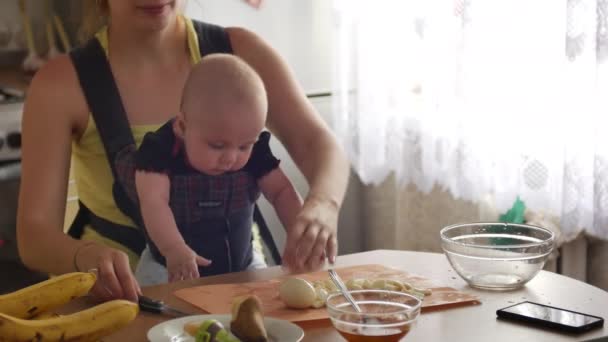 小さな男の子がスリングに座っている若い女性は、台所のテーブルに座っている間に卵をカットし、仕上げ後、彼女の子供にキスをします。母親の家事の概念。スローモーションフロントビューで4kビデオ — ストック動画