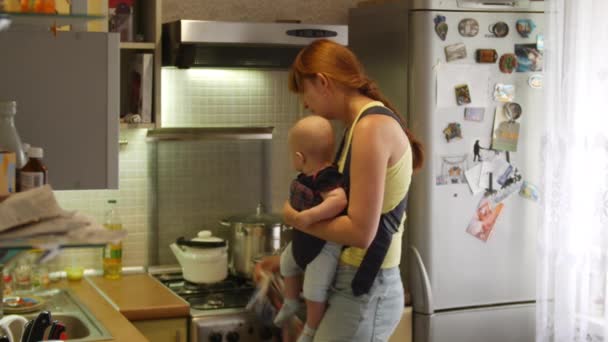 Kobieta z małym chłopcem siedzącym w temblaku gotuje zupę na obiad, sprząta w kuchni i całuje dzieciaka. Koncepcja obowiązków domowych matki. Widok z przodu w zwolnionym tempie w wideo 4k. — Wideo stockowe