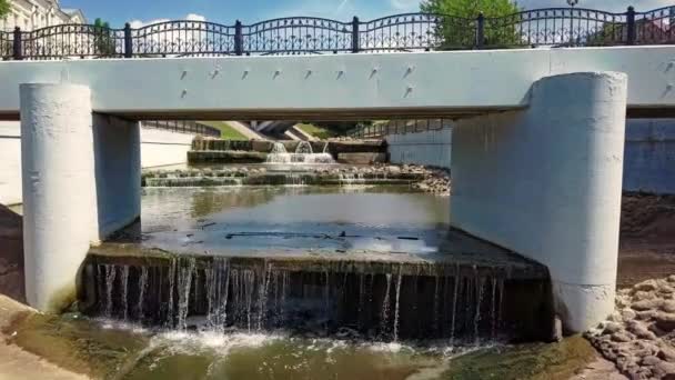 Drone voa até a ponte pedonal sobre um pequeno rio da cidade com um fluxo rápido e várias bordas que formam pequenas cachoeiras. Filme de câmera lenta vista aérea em vídeo 4K . — Vídeo de Stock