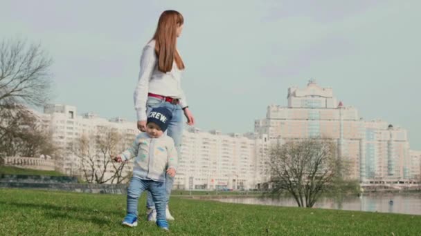 Menino e sua jovem mãe caminhando para a câmera no fundo da cidade primavera e alcançando a câmera o bebê sorri nele em vídeo de tiro médio 4K . — Vídeo de Stock