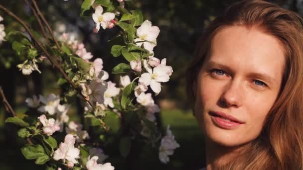 Mulher bonita apreciando a natureza no pomar de maçã primavera. Retrato de menina feliz no jardim com árvore de maçã florescendo no fundo. Modelo de moda sorridente perto de flores de flor. câmera lenta 4K vídeo — Vídeo de Stock