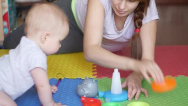 La mujer enseña a su bebé a montar una pirámide de juguetes jugando en el suelo sobre una alfombra colorida. Familia feliz y concepto de enseñar a los niños. Vista frontal plano medio en vídeo 4K — Vídeos de Stock