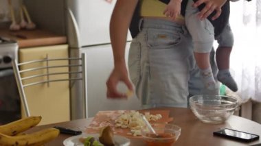 Yanında yeni yürümeye başlamış bir çocukla genç bir kadın mutfakta yemek için çorba pişiriyor. Anne ev işleri ve bebek bakıcılığı konsepti. 4k videosunda ön görünüm ortası görüntüsü