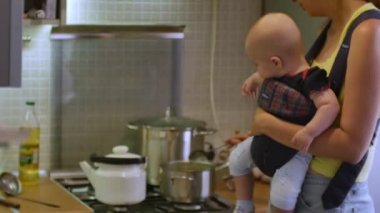 Bebek arabasında oturan bir çocukla güzel bir kadın mutfakta akşam yemeği için çorba pişiriyor. Anne ev işleri ve bebek bakıcılığı konsepti. 4k videosunda ön görünüm ortası görüntüsü.