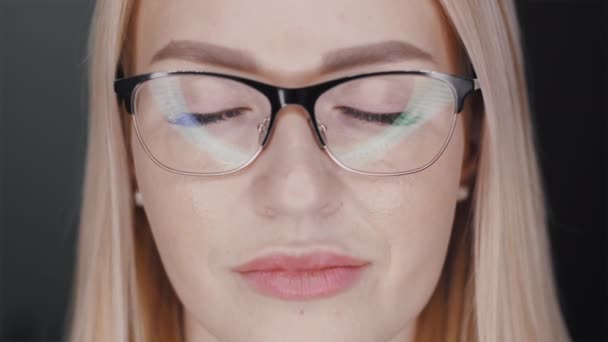 Portrait d'une jeune femme blonde et ambitieuse aux lunettes aux yeux fermés, puis elle les ouvre et regarde sérieusement à la caméra sur fond noir. Slow motion close up vue de face vidéo en 4K . — Video