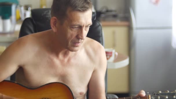 Staruszek gra na gitarze akustycznej z sześcioma strunami, siedzącej na krześle w kuchni. Widok z przodu średni strzał w 4k wideo. — Wideo stockowe