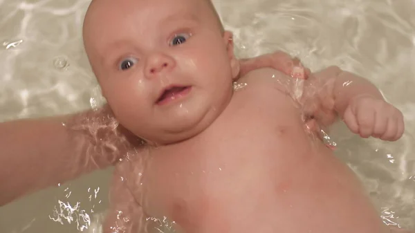 Újszülött baba fürdik csillogó vízzel a háttérben. A babát anyai kezek támogatják. Gyermek- és higiéniai koncepció. Közelkép portré lövés felső nézet 4k videó. — Stock Fotó