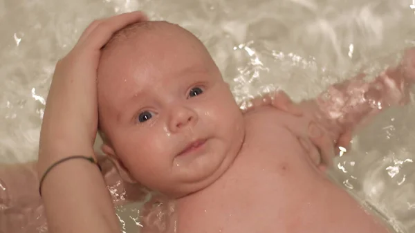 Bebé recién nacido tomando un baño con chispa de agua en el fondo. El bebé es apoyado por las manos de la madre. Concepto de niños e higiene. Vista superior de primer plano retrato en vídeo 4K . — Foto de Stock