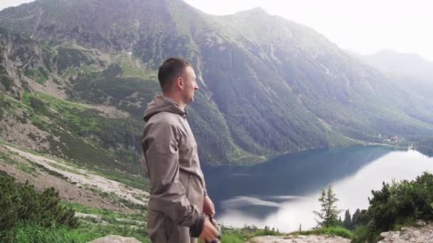 Seorang pemuda membuat foto panorama dari pemandangan indah dengan danau pegunungan dan puncak gunung hijau di latar belakang. Travel and adventure konsep slow motion side view 4K video. — Stok Video