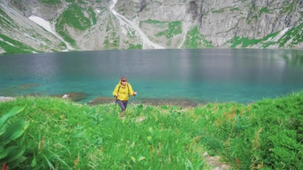 Una joven turista camina arriba con una caminata nórdica cuesta arriba desde un hermoso lago de montaña y disfruta de su aventura. Actividad recreativa deportiva. Turismo de vacaciones de verano. Vídeo en cámara lenta 4K . — Vídeo de stock