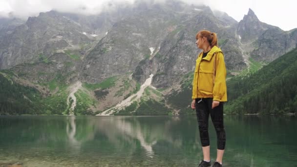 A tourist woman is standing on a stone at the shore of a beautiful mountain lake and looking at mountain peaks and then smiles into camera. Travel and adventure concept slow motion front view 4K video — Stock Video