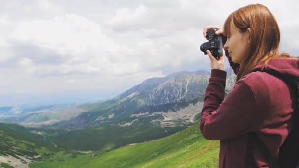Une jeune femme avec un sac à dos bleu fait des photos d'un beau paysage de vallée de montagne sur un appareil photo numérique dans une journée ensoleillée d'été. Concept de voyage et de blogging au ralenti back view vidéo 4K . — Video