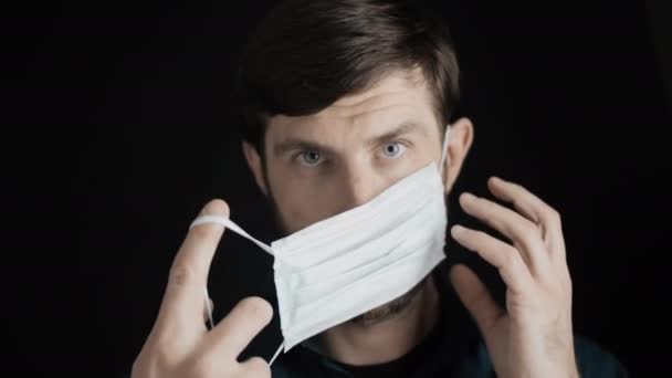 Aantrekkelijke man met een baard zet een chirurgisch masker op zijn gezicht voor corona virus covid-19 preventie. Infectiebescherming, quarantaine en epidemie. Langzame beweging close-up vooraanzicht 4k video. — Stockvideo