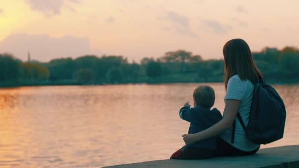 Mamá e hijo se sientan junto al agua y observan el atardecer — Vídeos de Stock