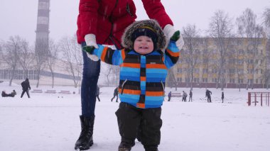 Anne, parktaki karda gülümseyen küçük bir bebeğin ellerine önderlik ediyor. Çocuğun ilk adımları. Ebeveyn bakımı
