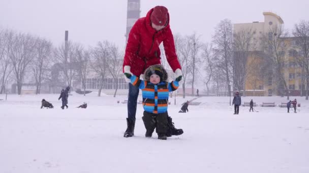 Мать ведет руки маленького улыбающегося ребенка в снегу в парке. Первые шаги малыша. Родительский уход — стоковое видео