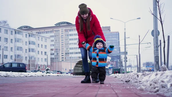 Una giovane donna conduce un bambino per le mani lungo la strada in inverno. Primi passi bambino — Foto Stock