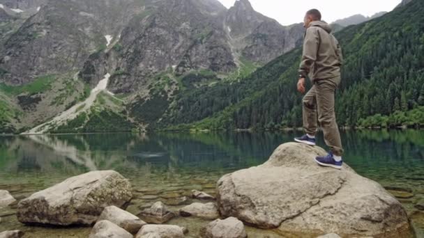 Touriste grimpe un rocher près d'un lac de montagne, photographie le paysage autour. Le concept de voyage et d'aventure. Mouvement lent — Video