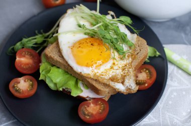 Kızarmış yumurtalı sandviç, et, domates, bezelye filizi ve salata. Siyah tabakta kahvaltı..