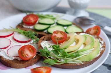 Krem peynirli vejetaryen sandviçler, salatalık, avokado, turp, domates ve bezelye filizleri beyaz tabakta. Beyaz stantta yumurta sisi. Vejetaryen yemekleriyle kahvaltı.