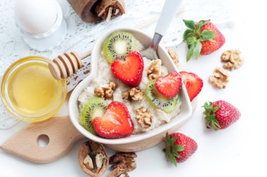Beyaz arka planda yulaf ezmeli kahvaltı, çilekli kivi, bal, ceviz ve yumurta. Çocuk için kahvaltı. Beyaz kalp şeklinde bir tabakta yulaf ezmesi. Üst görünüm