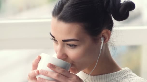 Okouzlující mladá žena v sluchátka sedí u okna poslech musik a pití kávy — Stock video