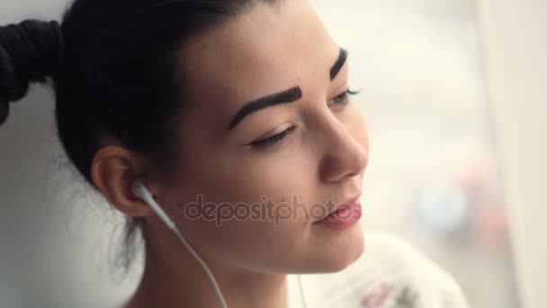 Charming young woman in earphones sitting near window listening musik — Stock Video
