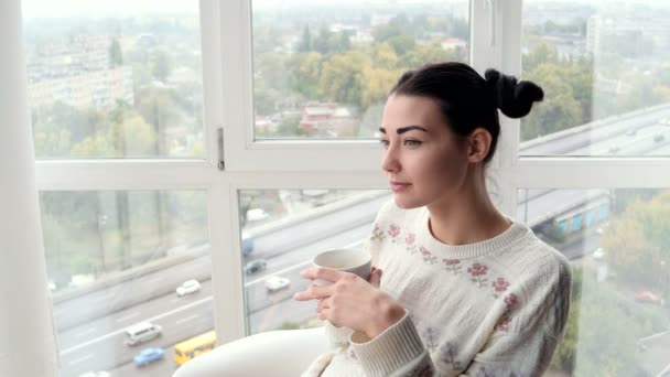 Charmante jonge vrouw zitten in de buurt van venster en drinken koffie — Stockvideo