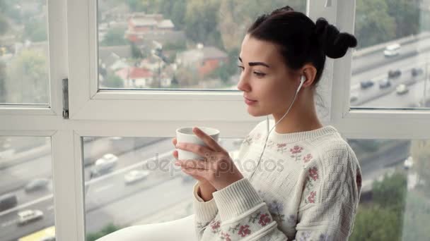 Очаровательная молодая женщина в наушниках сидит у окна, слушает музыку и пьет кофе — стоковое видео