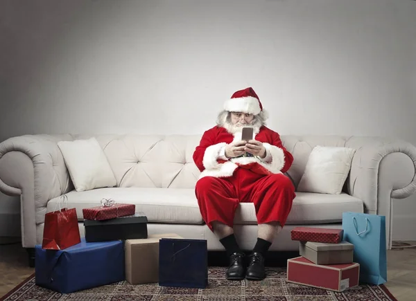 圣诞老人坐在沙发上 在他手中的智能手机和一些礼物周围 — 图库照片