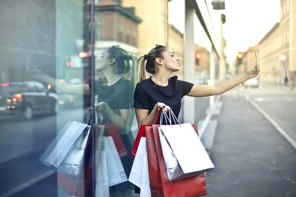 年轻妇女与一些购物袋采取自拍在商店窗口前面 — 图库照片