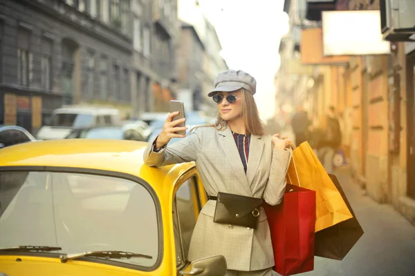 街で黄色のタクシーでサポートされている彼女の携帯電話を使用していくつかのショッピング バッグを持つエレガントな女性 — ストック写真
