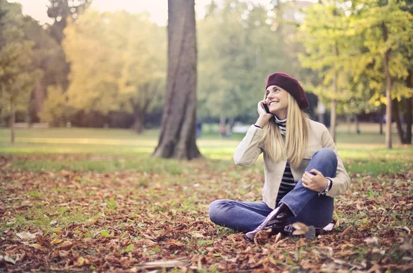 公園の地面に座って携帯電話で話している金髪の女性 — ストック写真